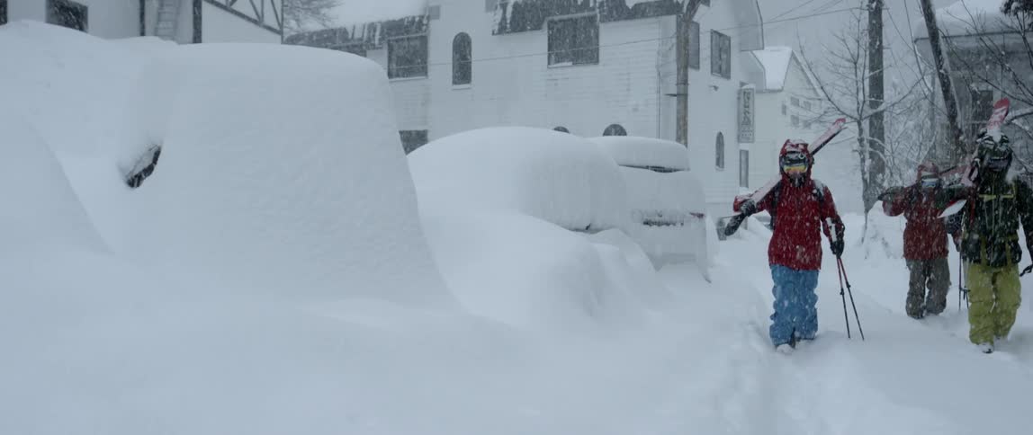 Three people with skiing equipment walk past snow covered cars