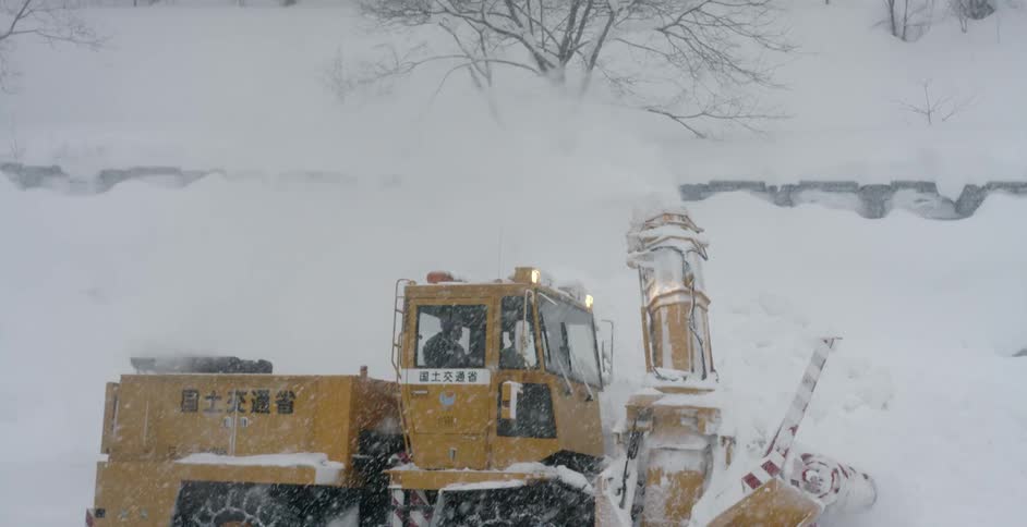 A snow plough with characters of an asian language emblazoned on its side attempts to clear a path under heavy snowfall