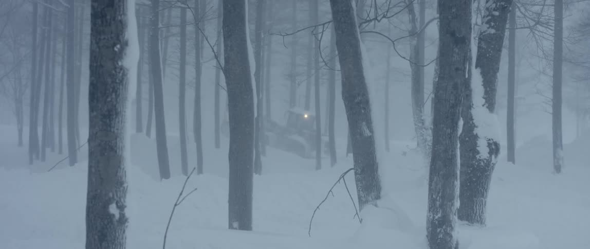A vehicle with lights moves through snowy woods