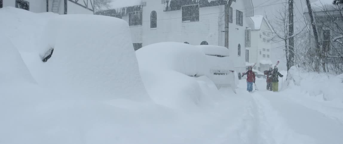 Three people with ski gear walk down a snow covered road past vehicles buried in snow