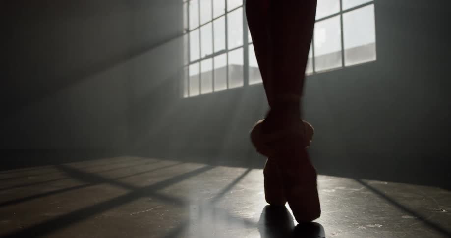 On her tippy toes a ballerina perfects her dance while wearing ballet shoes twirling around a dimly lit studio