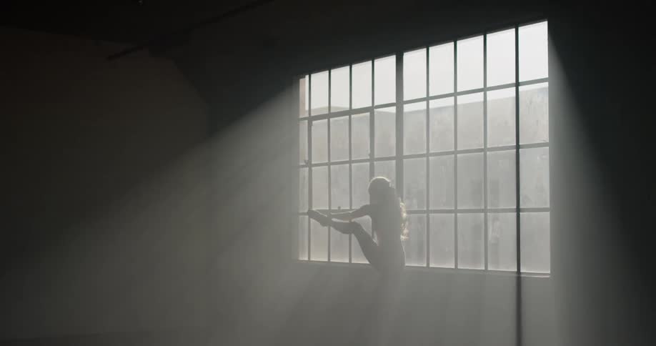 Ballet dancer holds leg up while standing near window in dark room