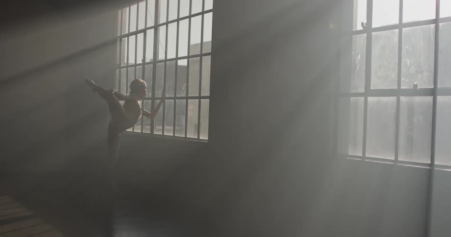 A dancer walks to a window in a dance studio and lefts and stretches her leg