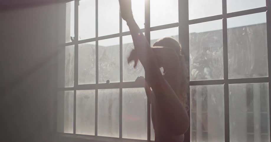 Young ballerinas silhouette dances in the dark room in front of the window while the sun shines