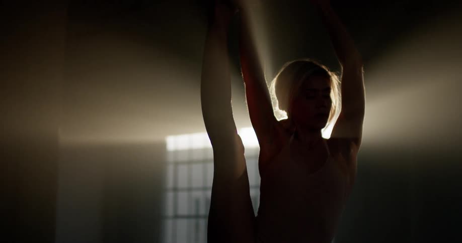 Ballet dancer rehearses in dark room backlit by sun in window