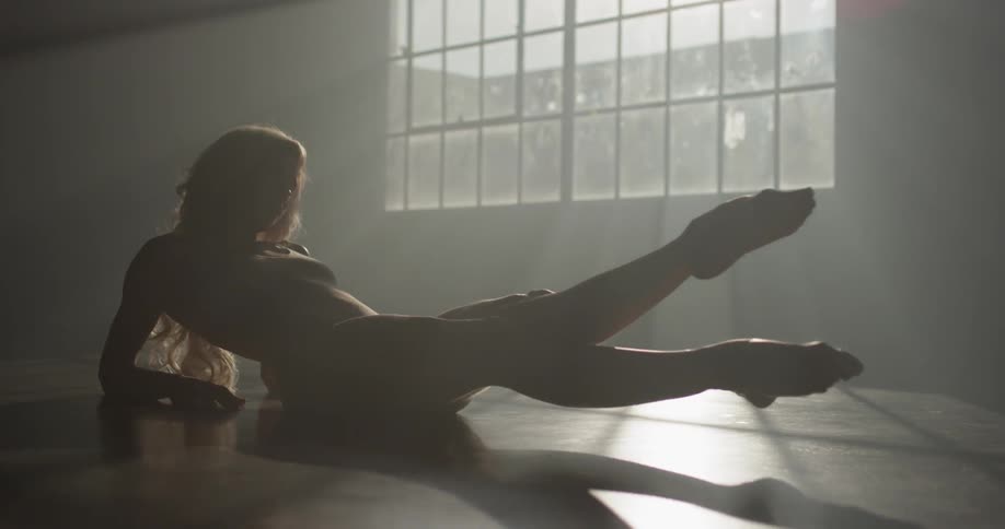 A white woman lies on the floor of a dance studio and stretches her legs