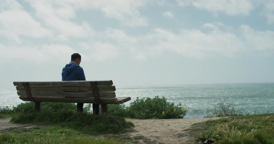 A man sits alone on a bench on a beach overlooking an ocean