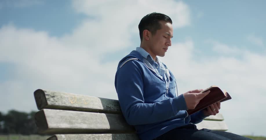 An asian man sits on a wooden bench and reads a book