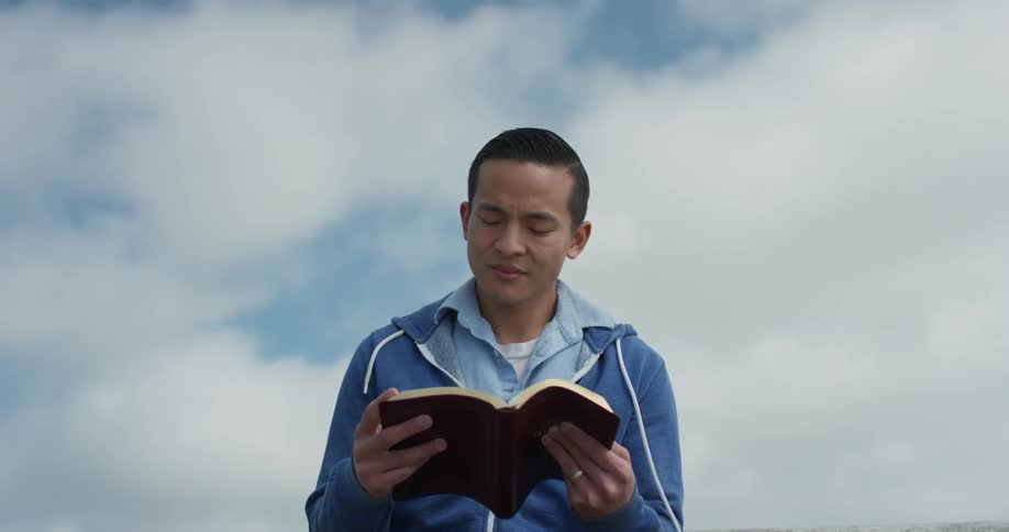An asian man reading a book under a cloudy blue sky