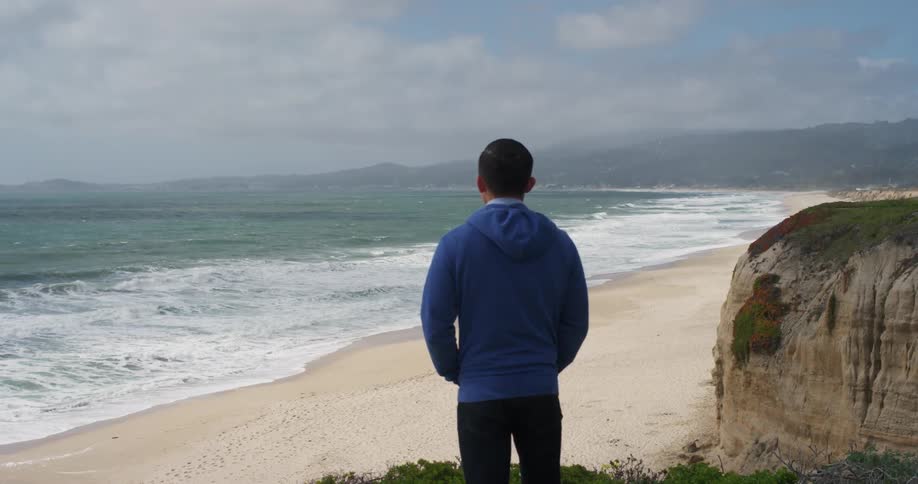 Overlooking the beach a young man is standing near a cliff as he looks over the incoming waves