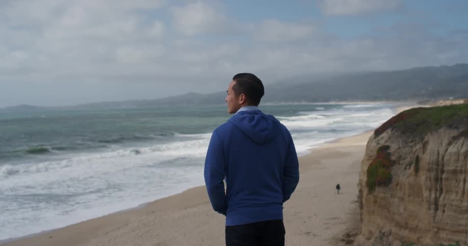A man stands near a cliff above beach as he gazes over the ocean as a man on the beach walks around with a metal detector trying to find some treasure