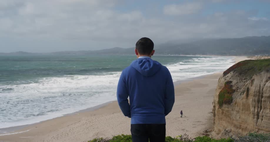 A man standing on a cliff looking out at the beach and ocean below