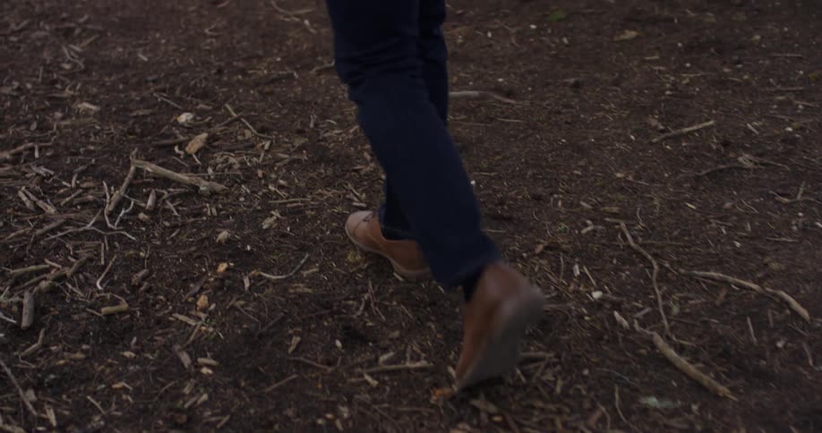 A person in brown shoes walks along a ground of bark and dirt