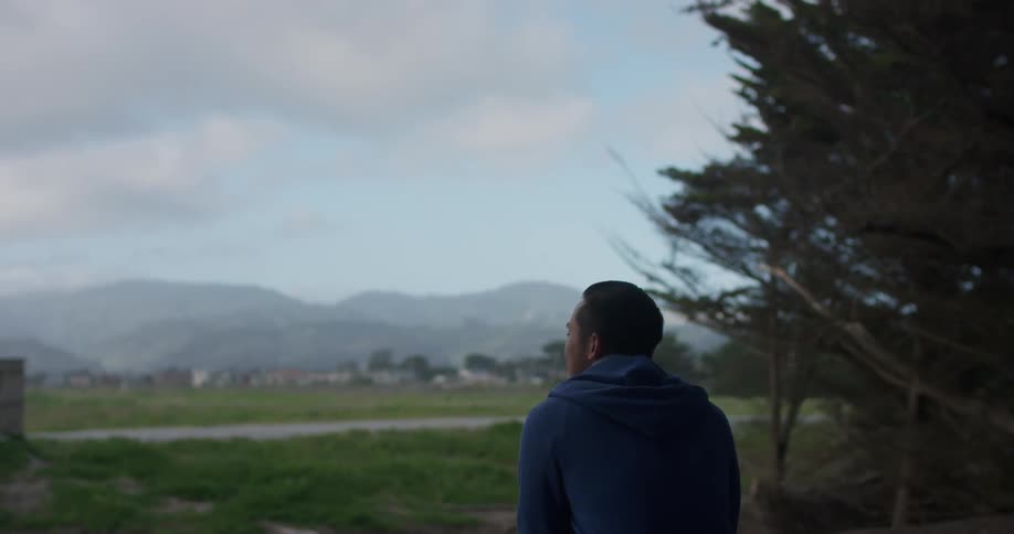 A man wearing a hoodie walks out of the trees and sits down and looks at the field and hills on the horizon