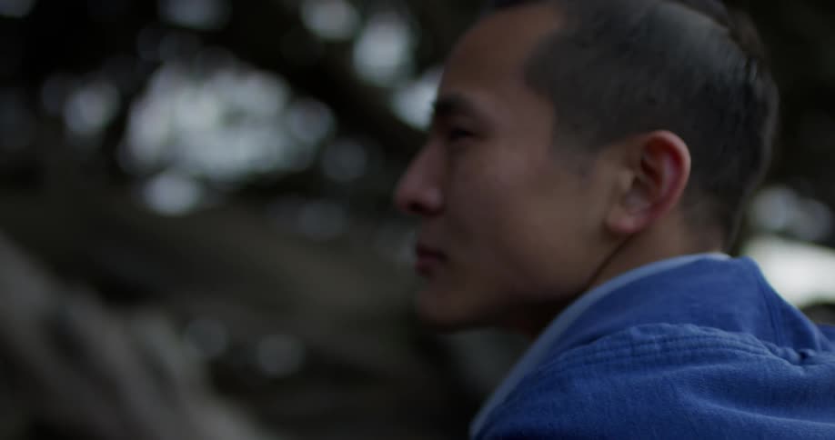 An asian man stands in front of large trees and looks around