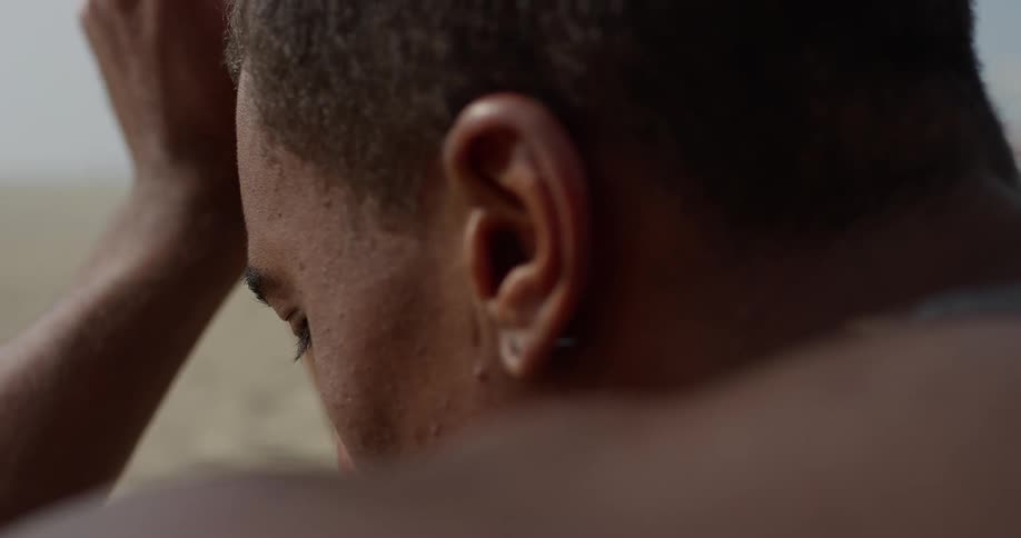 A black man sits on the beach and twists his hands in worry