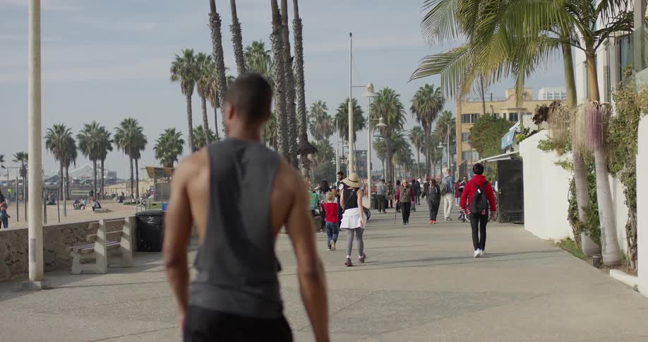 A muscular man walks down a sidewalk lined with palm trees near the beach