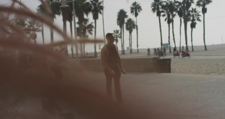 Black man wearingblack tank top walks on the concrete ground next to the beach with people on the background during daylight