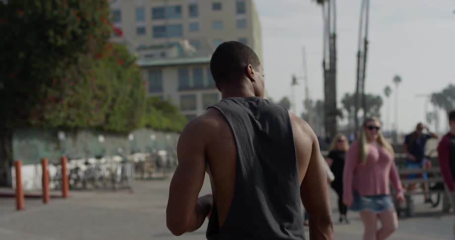 Black man wearing tank top walks on the concrete ground with people on the background next to the beach during daylight