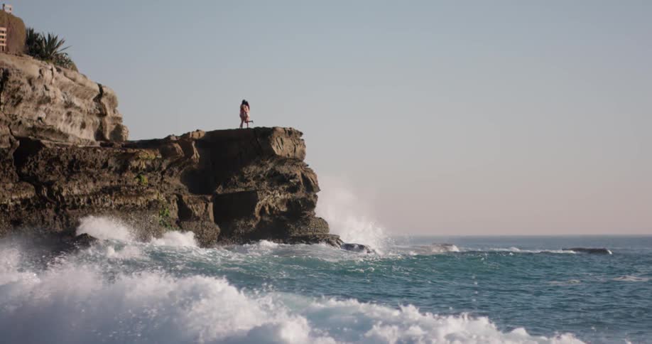 Ocean waves crash into the base of a cliff with two people standing above