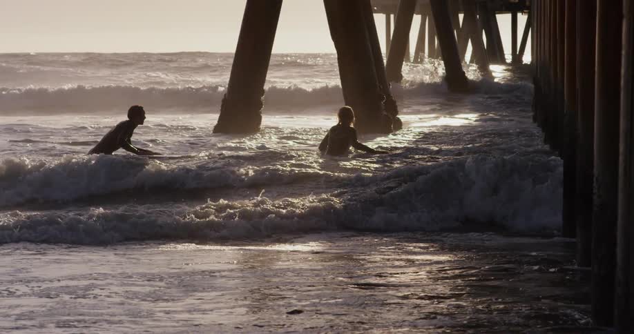 Two surfers wade in ocean waves beneath a pier at sunset