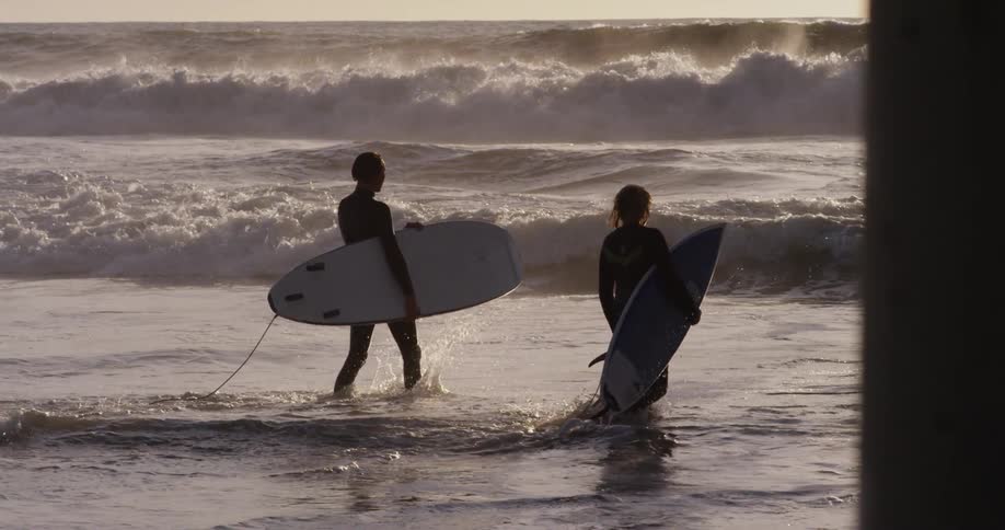 A man and woman walk through the water carrying surf boards