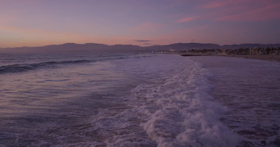 A bird flying over waves crashing on a beach at sunset with mountains in the background