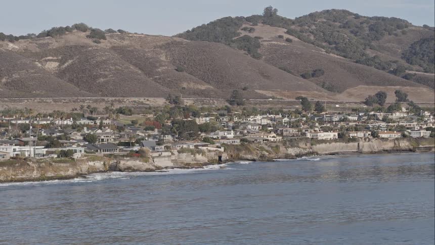 Waves splash on the shore of a coastal neighborhood while cars travel on roads at the base of the hills