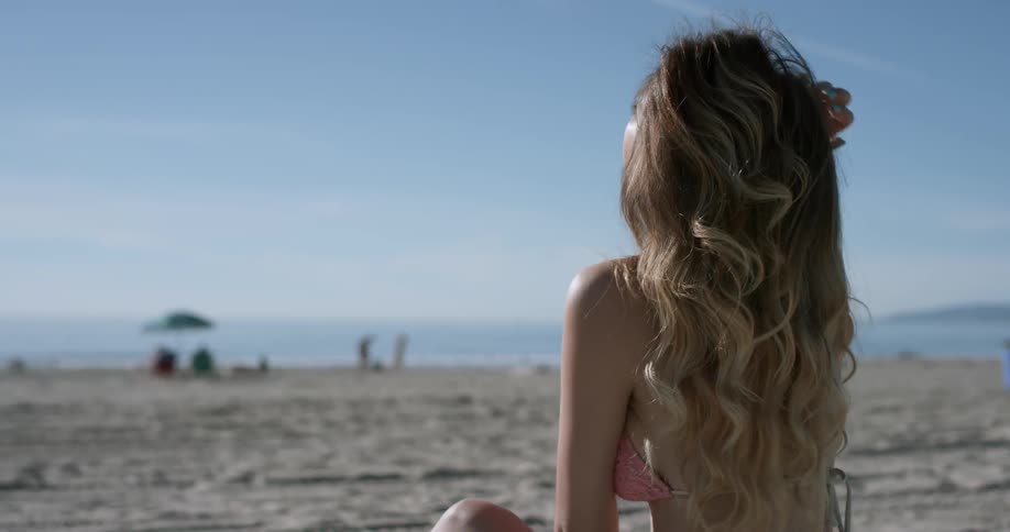 A woman wearing a bikini and sitting on the beach runs her hand through her long hair