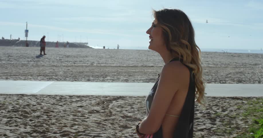 A white woman wearing a bikini top covered with a shirt walks along a beach and smiles