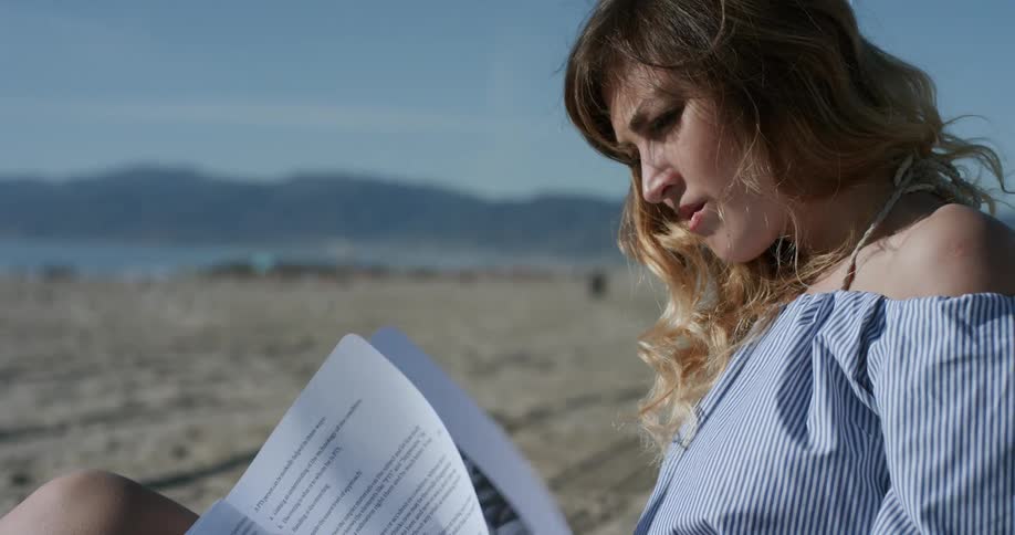 A woman in a blue striped dress reads a book on a sandy beach