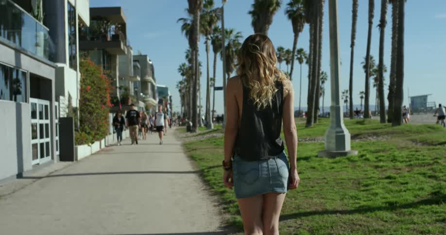 A white woman in a short denim skirt walks on a sidewalk down at the beach