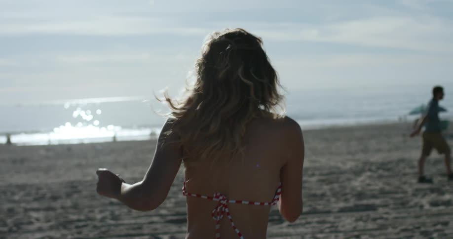 A woman wearing a bikini is running on the beach towards the ocean and then turns around laughing