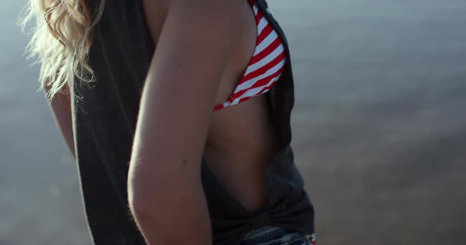 A young woman stand near water wearing a bikini top jeans and a tank top