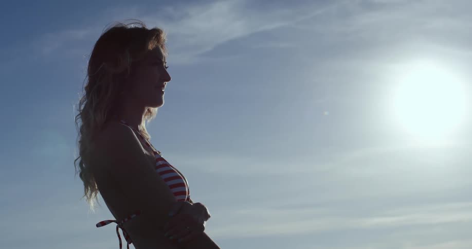 A woman in a bikini stands outside and grabs right arm with her left arm