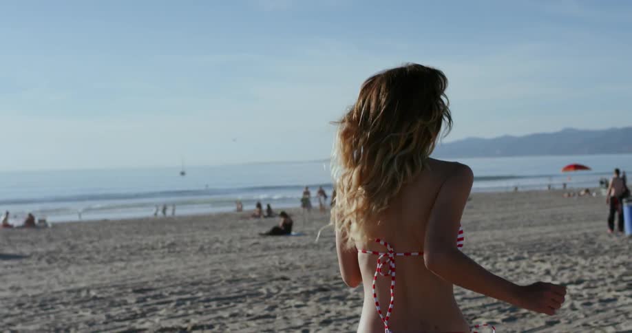 A woman jogs across the sandy beach toward the water and turning to laugh behind her