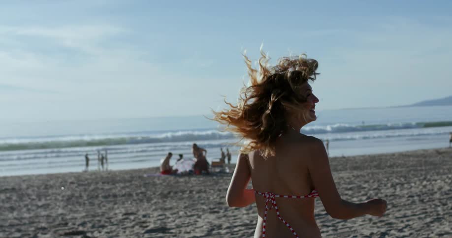 A young woman in a bikini runs on the beach before turning around laughing
