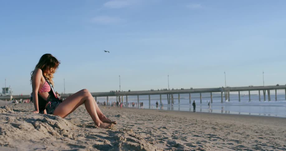 Woman wearing mini short sits on a sand dune on the beach and plays sand with feet next to the sea