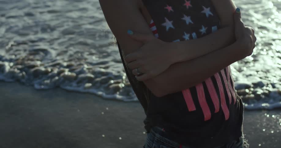 A girl stands on the beach next to the water and runs her hands along her upper arms