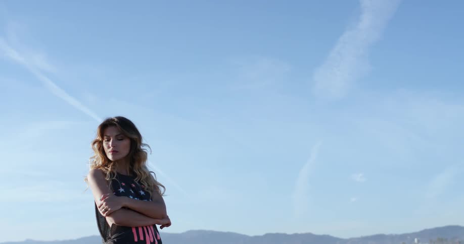 A woman in front of a mountainous backdrop looks out towards the sun and preens
