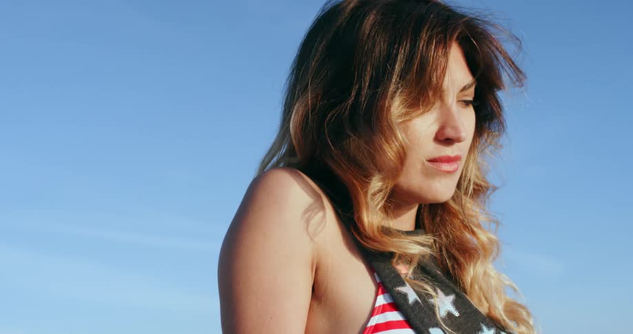 A white woman wearing a bikini top and a loose shirt sits on the beach with her eyes closed and then stands up