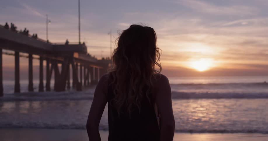 Woman stands on a beach next to a pier looking at sunset
