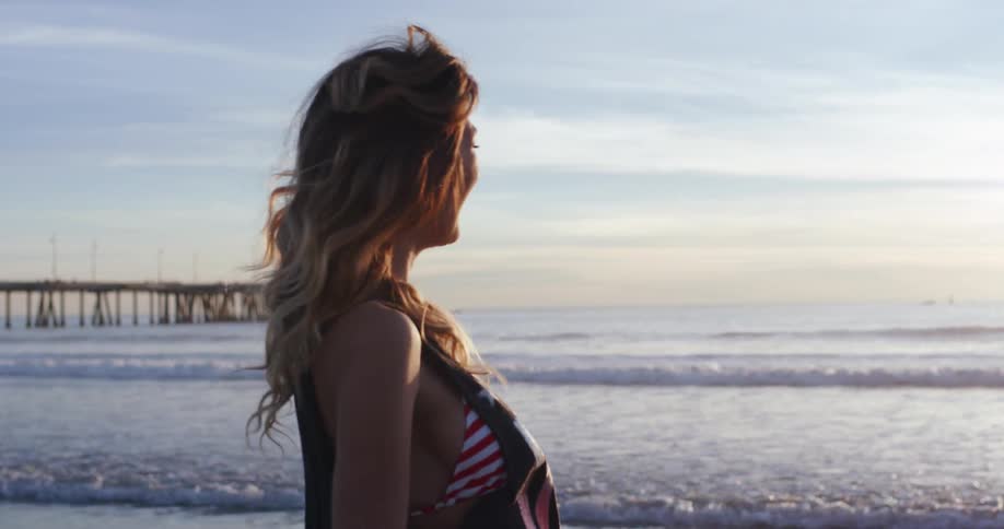 A woman throws sand while walking barefoot on a beach at sunset