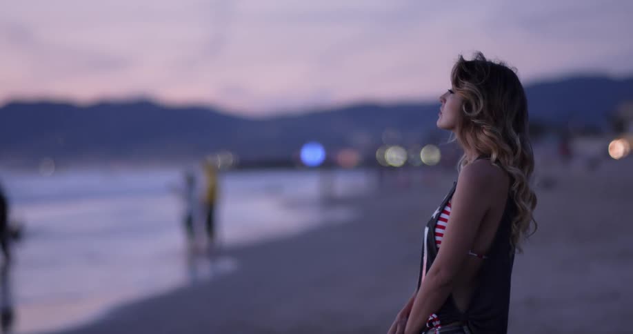 A young woman stands on the beach looking out at the ocean at sunset