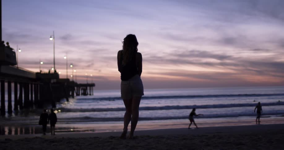 A woman looks out onto the ocean while standing on a beach and a couple play frisbee in the sand in front of her
