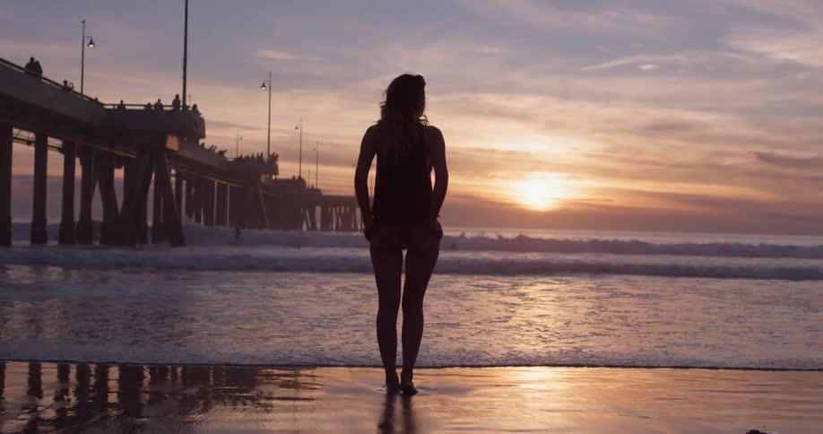 Woman stands on a beach next to a pier in front of the sunset over the ocean