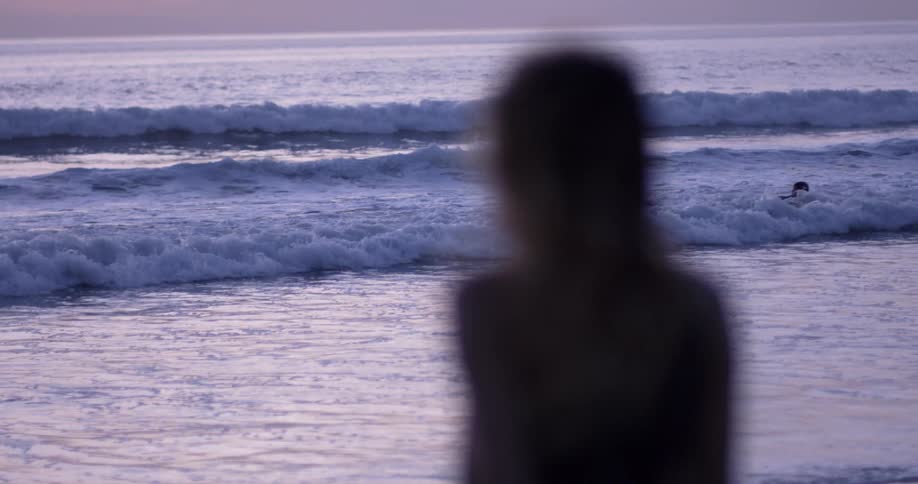 A woman sits watching two children play in the waves on a beach at dusk