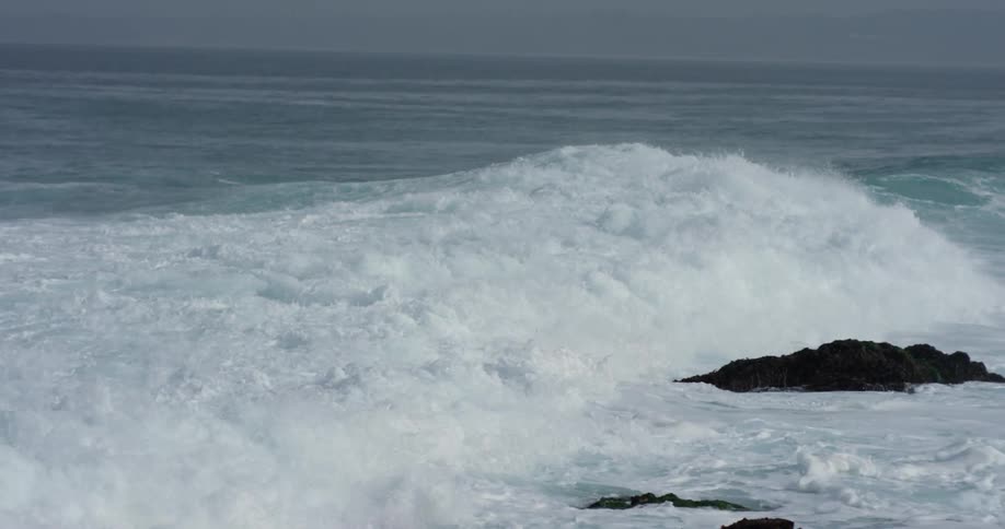 Two seagulls fly over waves crashing against the shore during daylight