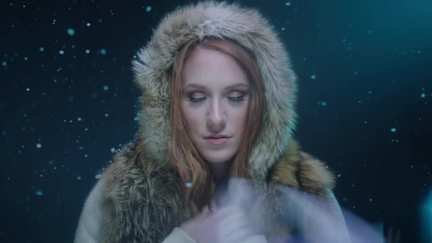 A young woman wearing a hooded coat stands in the snow with her eyes clothes