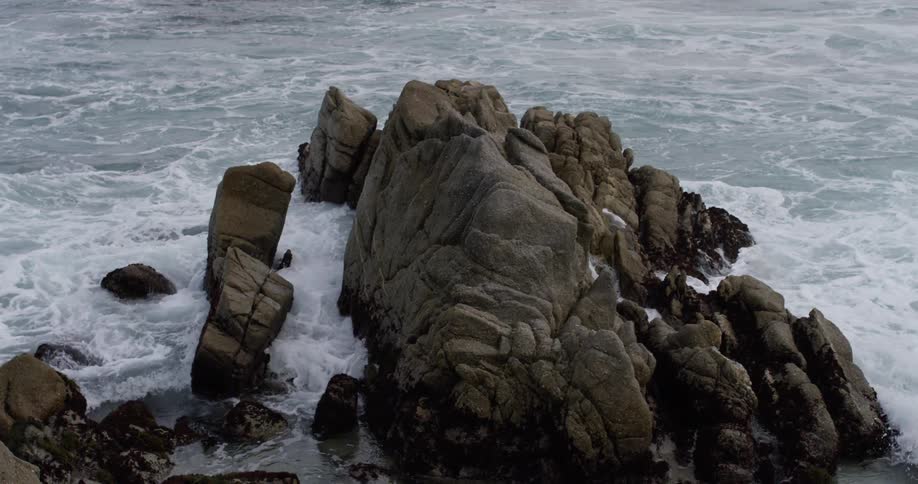 Waves crash in slow motion against large rocks in ocean